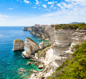 Kalksteinfelsen und t&uuml;rkisblaues Meer vor der Stadt Bonifacio auf Korsika, mit dramatischer Steilk&uuml;ste, &uuml;ppiger Vegetation und klarer Sicht auf das Mittelmeer unter einem teils bew&ouml;lkten Himmel.