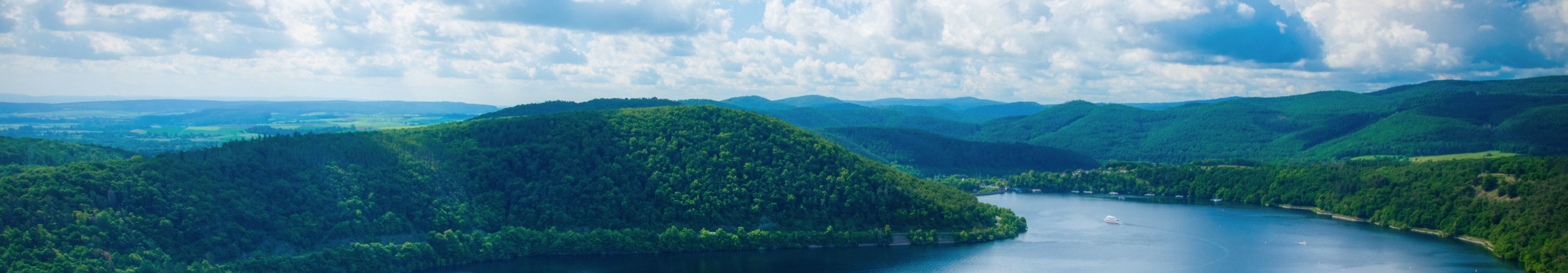 Panoramablick auf den Edersee im Nationalpark Kellerwald-Edersee in Hessen, umgeben von bewaldeten H&uuml;geln unter blauem Himmel mit wei&szlig;en Wolken.