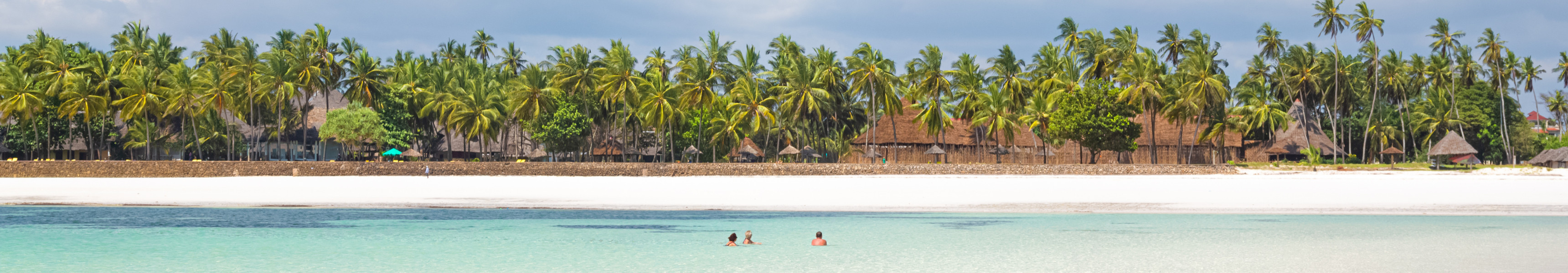 Wei&szlig;er Sandstrand mit t&uuml;rkisfarbenem Wasser und badenden Menschen am Diani Beach in Kenia, umgeben von Palmen und traditionellen Strohh&uuml;tten.