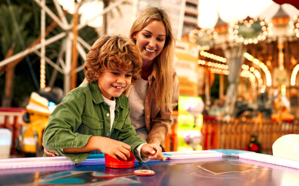 Mutter und Sohn spielen zusammen an einem Airhockey-Tisch im Bayern Park, umgeben von bunten Lichtern und Fahrgesch&auml;ften.