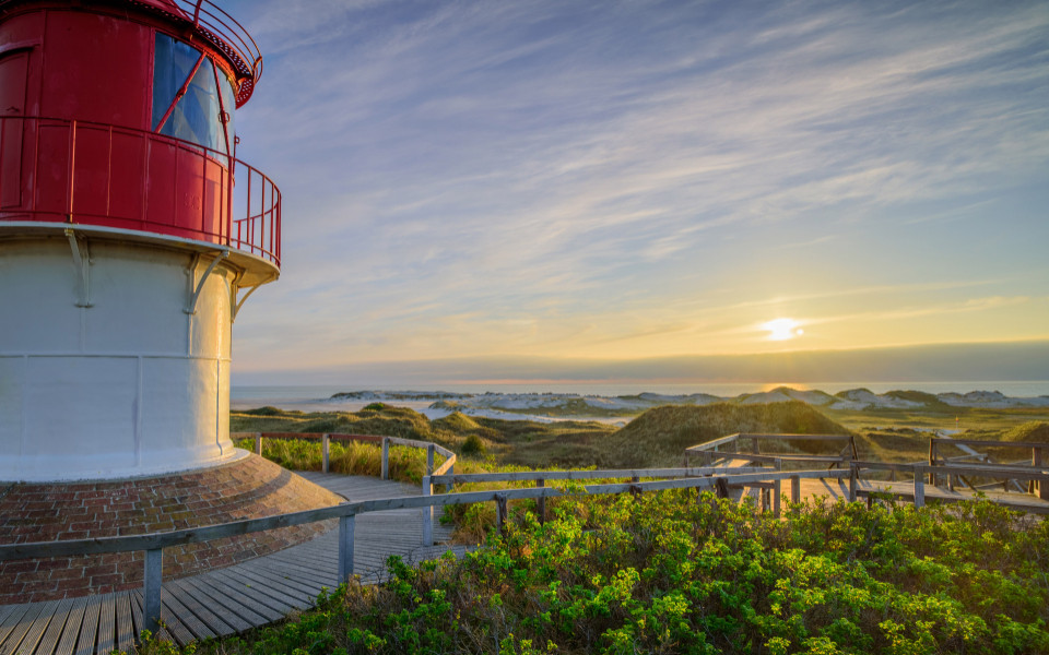 Der Leuchtturm auf der Insel Amrum, umgeben von gr&uuml;nen D&uuml;nen und dem weiten Blick auf die Nordsee bei Sonnenuntergang.