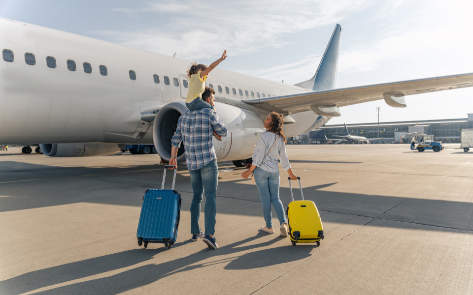Familie läuft mit Koffern zum Flugzeug und das Kind sitzt auf den Schultern des Papas.