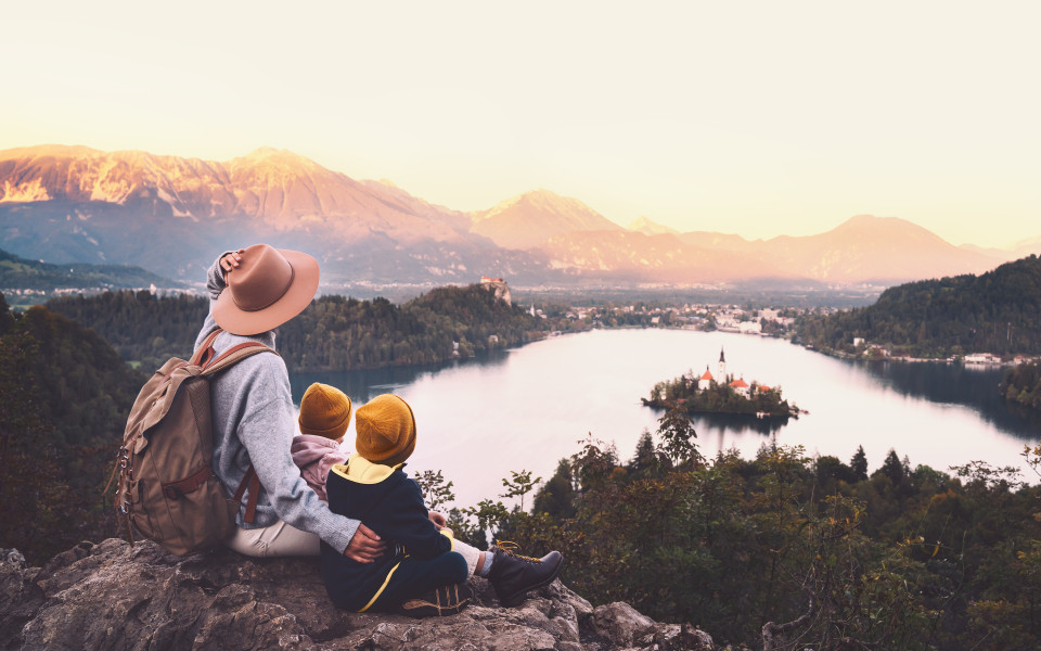 Mutter mit zwei kleinen Kindern sitzt auf einem Felsen und blickt auf den Bleder See in Slowenien – mit der Inselkirche im Wasser und schneebedeckten Bergen im Hintergrund bei Sonnenuntergang.